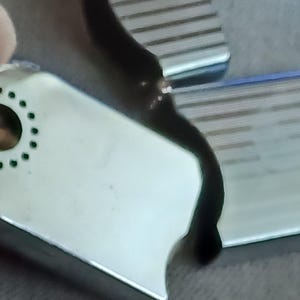 May include: A white rectangular tag with a circular hole surrounded by a ring of small green dots. The tag is positioned against a dark, curved surface with a metallic, ribbed texture visible in the upper right corner of the frame.