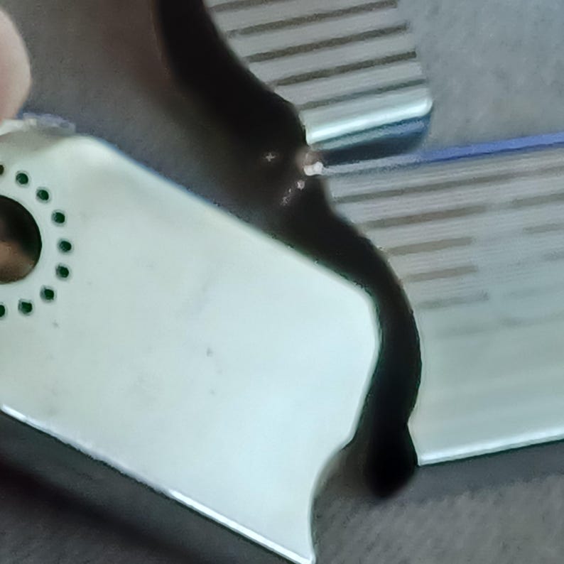 May include: A white rectangular tag with a circular hole surrounded by a ring of small green dots. The tag is positioned against a dark, curved surface with a metallic, ribbed texture visible in the upper right corner of the frame.