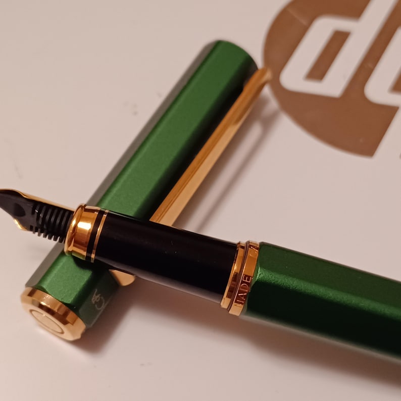 Puede incluir: Una pluma estilogr&aacute;fica verde con detalles dorados y una plumilla negra. La pluma tiene la palabra "MADE" grabada en el lateral.