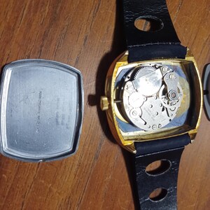 Puede incluir: Un reloj de pulsera vintage dorado con una correa de cuero negro. La esfera del reloj est&aacute; abierta, revelando los intrincados mecanismos internos. La parte trasera del reloj est&aacute; separada, mostrando la inscripci&oacute;n "AER CHROMATIC WATCH CO."