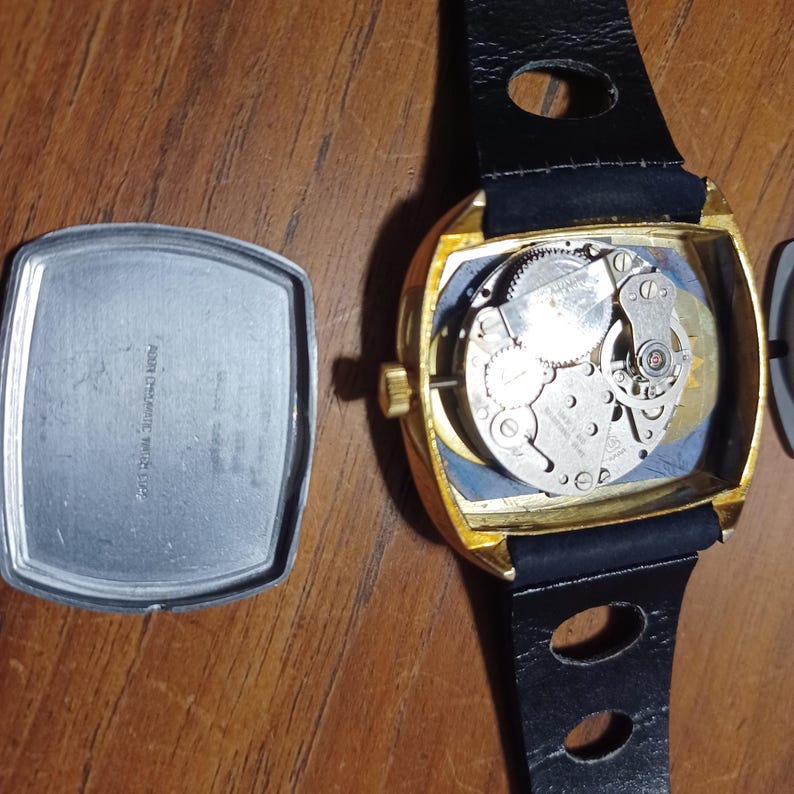 Puede incluir: Un reloj de pulsera vintage dorado con una correa de cuero negro. La esfera del reloj est&aacute; abierta, revelando los intrincados mecanismos internos. La parte trasera del reloj est&aacute; separada, mostrando la inscripci&oacute;n "AER CHROMATIC WATCH CO."