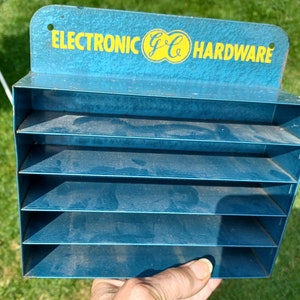 May include: A blue metal display case with seven shelves. The case has a sign that reads "ELECTRONIC G&C HARDWARE".