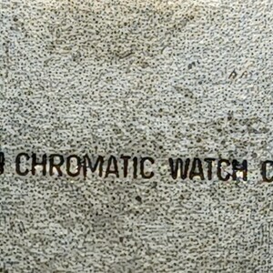 Puede incluir: Primer plano de una superficie texturizada y moteada con el texto "AGON CHROMATIC WATCH CORP" en color oscuro. El texto est&aacute; centrado y parece estar estampado o impreso en la superficie.
