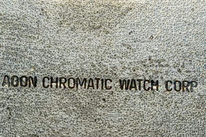 Puede incluir: Primer plano de una superficie texturizada y moteada con el texto "AGON CHROMATIC WATCH CORP" en color oscuro. El texto est&aacute; centrado y parece estar estampado o impreso en la superficie.