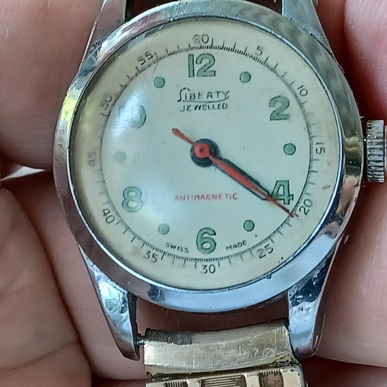 May include: Vintage Liberty watch with a silver-tone case and a cream-colored face. The watch features black numerals, a red second hand, and the text "Liberty Jeweled" and "Antimagnetic". The watch band is a gold-tone metal.