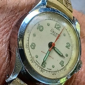 May include: Close-up of a vintage wristwatch with a cream-colored face. The watch features black numerals, a red second hand, and green hour and minute hands. The watch is marked "Swiss Made" and "Antimagnetic". The watch band is gold and silver.