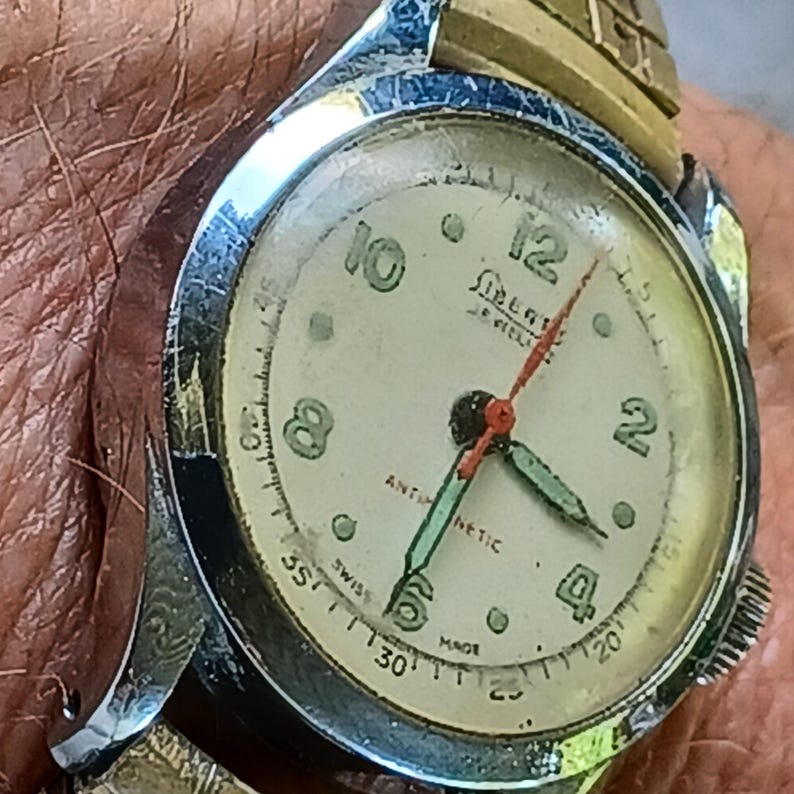 May include: Close-up of a vintage wristwatch with a cream-colored face. The watch features black numerals, a red second hand, and green hour and minute hands. The watch is marked "Swiss Made" and "Antimagnetic". The watch band is gold and silver.