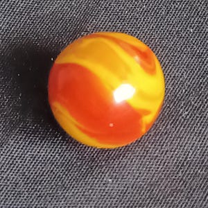 May include: A vibrant, swirled glass marble with a glossy finish. The marble features a combination of orange and yellow colors, creating a dynamic and eye-catching pattern. The spherical shape is clearly defined, making it a classic toy or collectible.