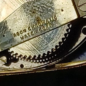 Puede incluir: Primer plano de un mecanismo de reloj con el texto "AGON CHROMATIC WATCH CORP." La imagen muestra engranajes intrincados y una superficie met&aacute;lica texturizada. El reloj parece ser vintage, con un dise&ntilde;o detallado.