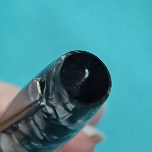 May include: Close-up of a vintage fountain pen nib. The pen features a black ink reservoir and a silver-colored clip. The pen's body has a marbled, gray and black pattern. The background is a solid teal color.
