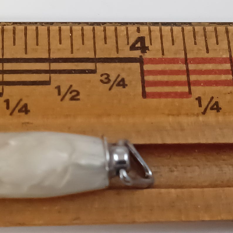 May include: Close-up of a small, pearly white pendant with a silver-coloured bail. The pendant is next to a wooden ruler, showing measurements. The pendant's design is simple and elegant.