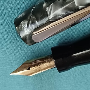 May include: Close-up of a vintage fountain pen with a gold nib and black barrel. The pen's cap is a marbled gray and white. The nib is engraved with text. The pen is on a light blue surface.