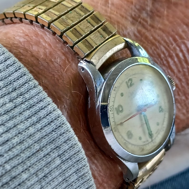 May include: Close-up of a vintage wristwatch with a gold-toned expanding band. The watch face is cream-colored with black numerals and hands. The watch case is silver-toned. The watch is on a wrist.