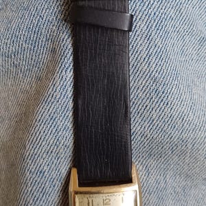 K&ouml;nnte beinhalten: Eine goldfarbene Bulova Duo Wind Armbanduhr mit einem schwarzen Lederarmband. Das Zifferblatt der Uhr ist wei&szlig; mit schwarzen r&ouml;mischen Ziffern als Stundenmarkierungen und schwarzen Zeigern.