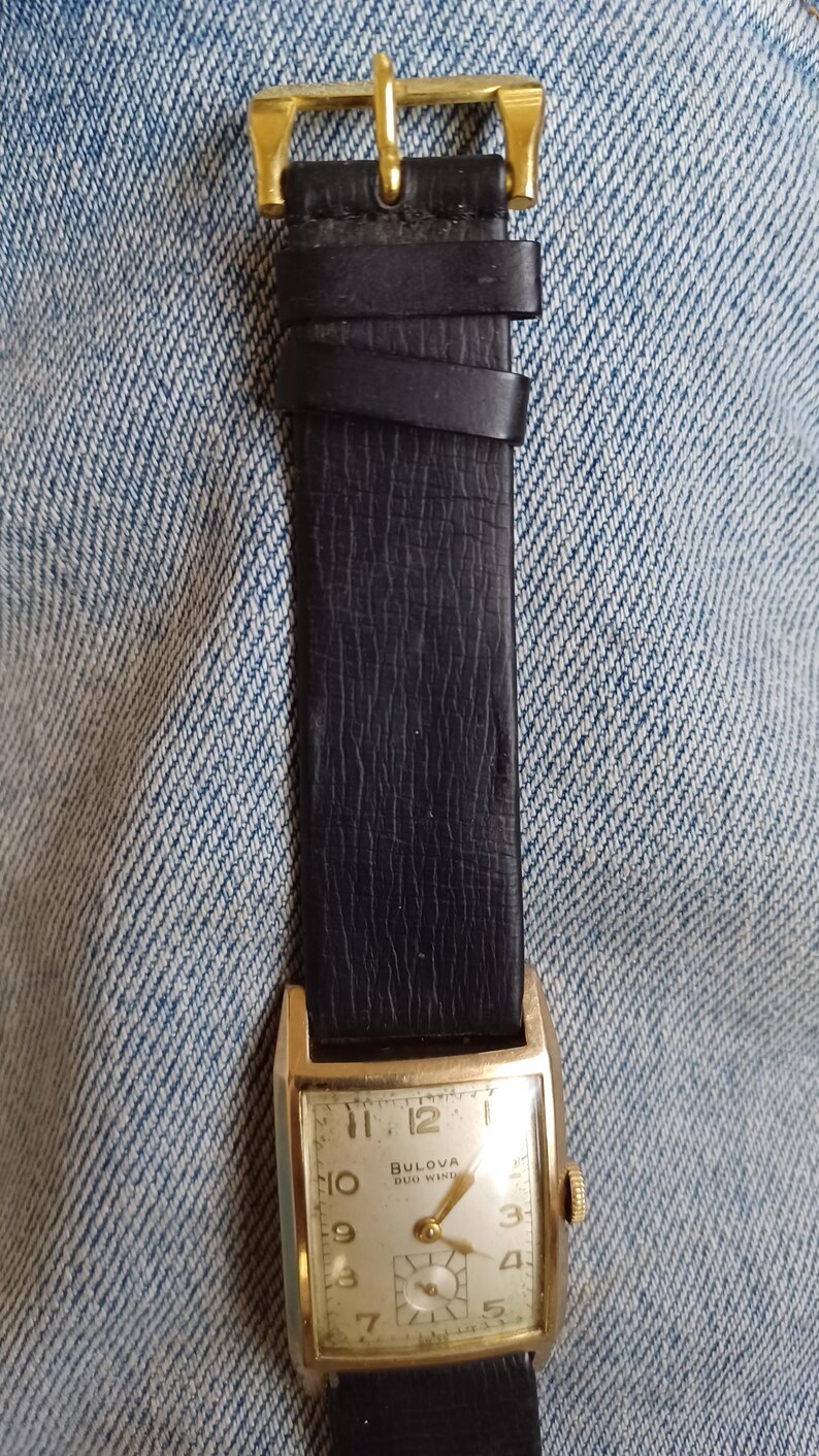 K&ouml;nnte beinhalten: Eine goldfarbene Bulova Duo Wind Armbanduhr mit einem schwarzen Lederarmband. Das Zifferblatt der Uhr ist wei&szlig; mit schwarzen r&ouml;mischen Ziffern als Stundenmarkierungen und schwarzen Zeigern.