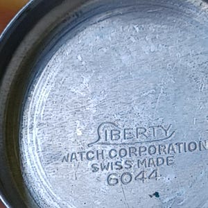 May include: Close-up of a watch back with the text "LIBERTY WATCH CORPORATION SWISS MADE 6044" etched into the metal. The watch back is a dull silver color with a textured surface.
