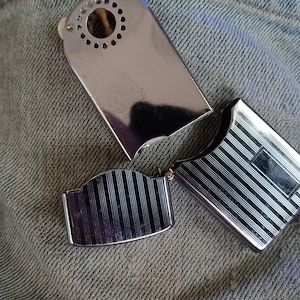 May include: A silver-toned metal lighter with a polished finish. The top section features a circular vent pattern, while the lower body and the hinged lid are decorated with a series of vertical engraved lines and a smooth rectangular panel.