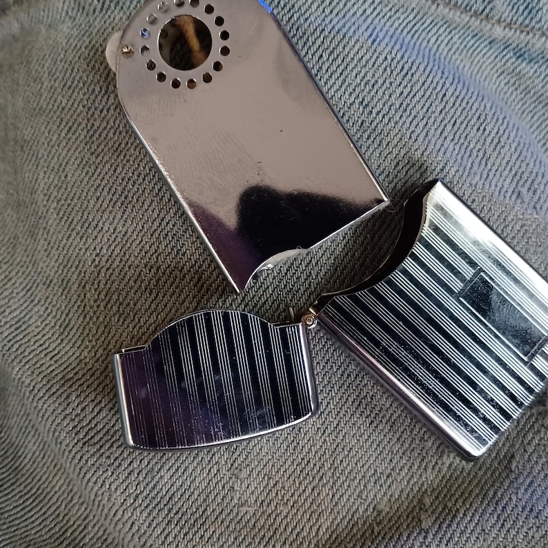 May include: A silver-toned metal lighter with a polished finish. The top section features a circular vent pattern, while the lower body and the hinged lid are decorated with a series of vertical engraved lines and a smooth rectangular panel.