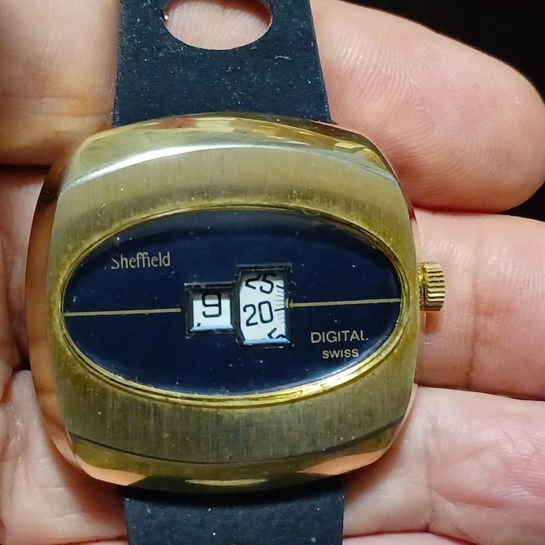 Puede incluir: Un reloj digital Sheffield vintage con caja dorada y correa negra. La esfera es azul oscuro con n&uacute;meros blancos que muestran la hora. Las palabras "Sheffield" y "DIGITAL SWISS" son visibles.