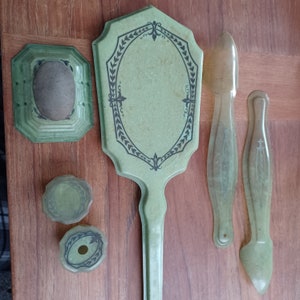 May include: A vintage green vanity set with a hand mirror, two hair combs, and two small round containers. The mirror has a decorative frame with a floral design.