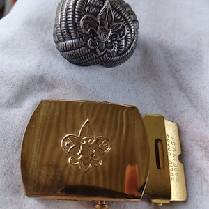 May include: A gold-colored belt buckle with a Boy Scout emblem and the words "Solid Brass Made in U.S.A."  A silver-colored Boy Scout emblem is also pictured.