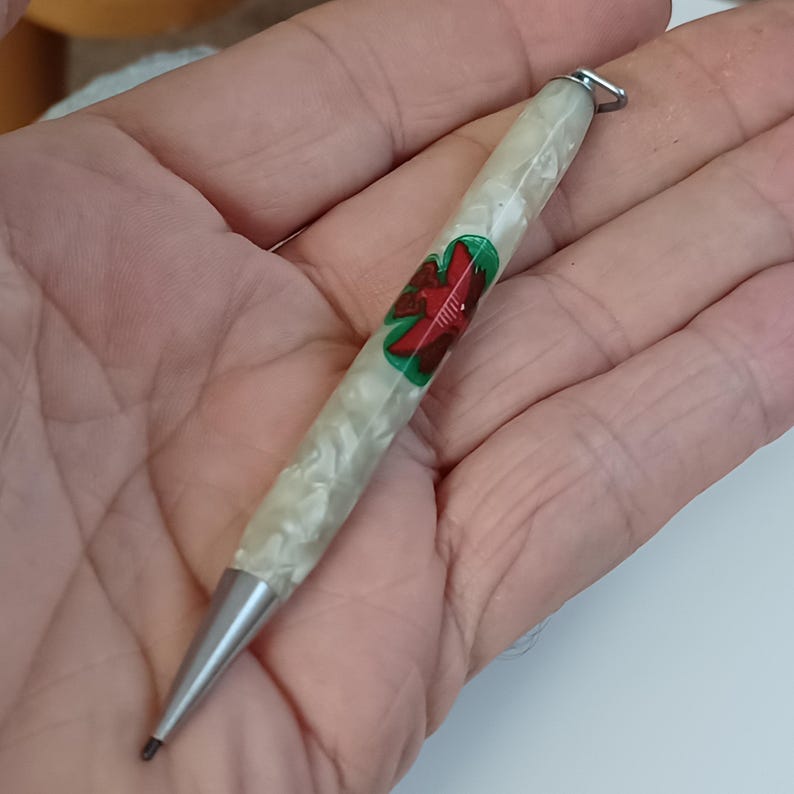 May include: A vintage mechanical pencil with a pearlescent white barrel and a silver tip. The barrel features a red and green floral design. A small silver loop is attached to the top of the pencil.