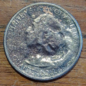 May include: A close-up of a United States quarter coin. The coin is silver in color and shows signs of wear. The words "LIBERTY", "QUARTER DOLLAR", and "UNITED STATES OF AMERICA" are visible.