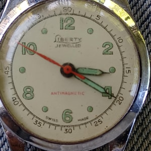 May include: Vintage Liberty watch with a cream-colored face, featuring black numerals and green accents. The watch has a red second hand and green hour and minute hands. The words "Liberty Jewelled" and "Antimagnetic" are visible on the face.
