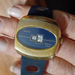 Puede incluir: Un reloj digital Sheffield vintage con caja dorada y esfera azul oscuro. El reloj muestra la hora 9:25. El reloj tiene una correa azul oscuro. La palabra "Sheffield" est&aacute; impresa en la esfera.