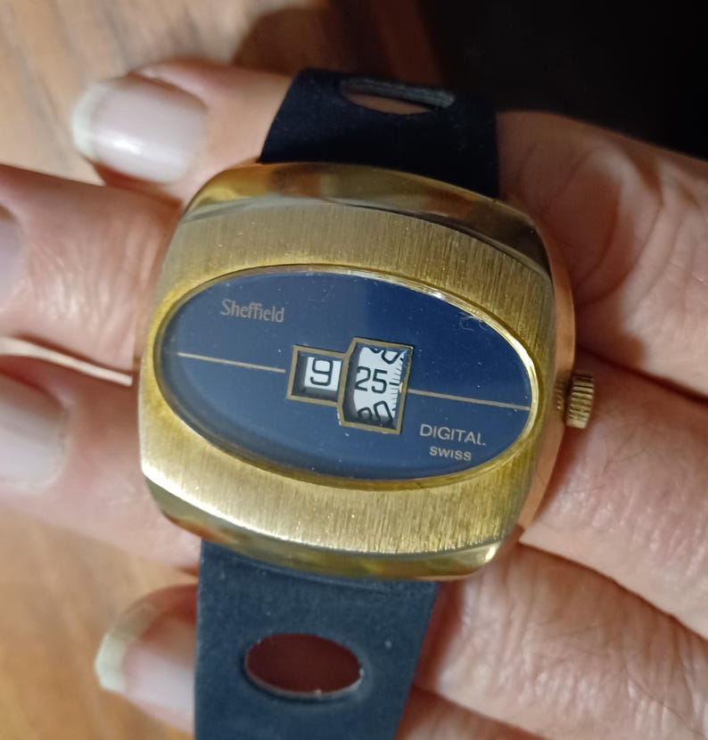 Puede incluir: Un reloj digital Sheffield vintage con caja dorada y esfera azul oscuro. El reloj muestra la hora 9:25. El reloj tiene una correa azul oscuro. La palabra "Sheffield" est&aacute; impresa en la esfera.
