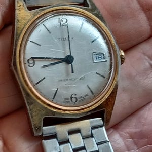 May include: Vintage Timex watch with a silver face, gold-tone bezel, and a silver metal band. The watch face displays the number 12, 9, 6, and 1, along with the date. The watch is marked "WATER RESISTANT" and "ENGLAND".