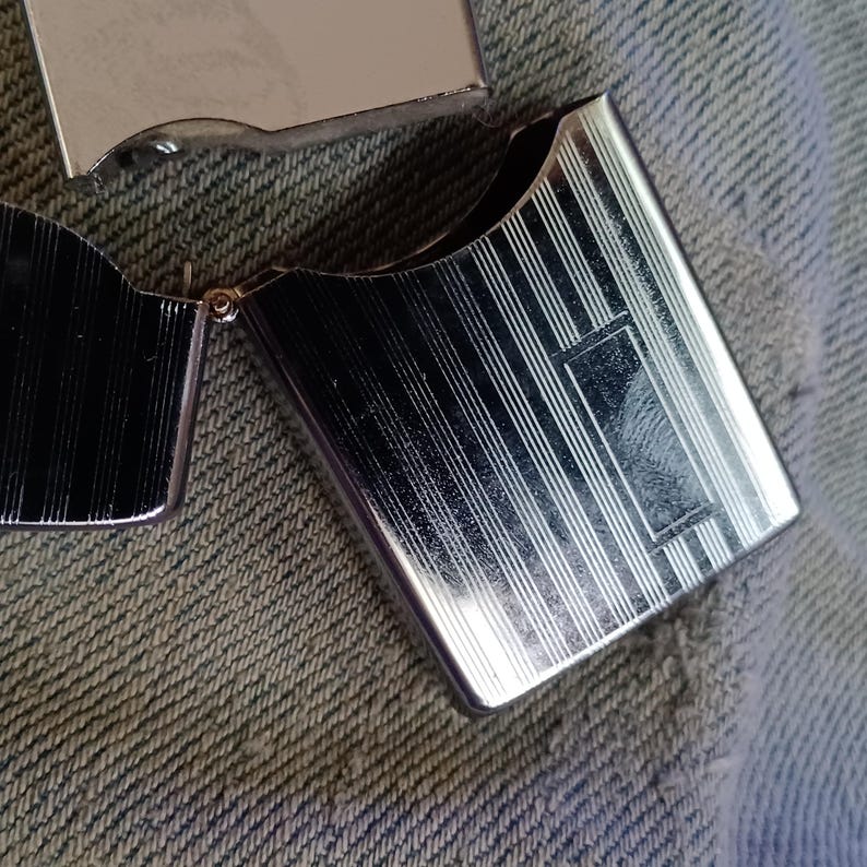 May include: A silver-toned metal lighter with a hinged lid, featuring a polished finish and a series of vertical engraved lines across the front surface. The lighter is shown in an open position, revealing the internal hinge mechanism and metallic casing.