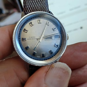 May include: A vintage Timex watch with a silver face, day and date display, and a brown leather band. The watch has a round silver case and a textured band. The watch face displays the word "TIMEX" and "WATER RESISTANT".