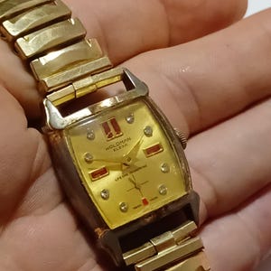 May include: A vintage gold-toned Woldman Elexa wristwatch. The watch features a rectangular gold face with diamond accents and red rectangular markers. The watch band is also gold-toned and has a classic link design. The watch face reads "WOLDMAN ELEXA" and "LIFETIME SPRINGING".