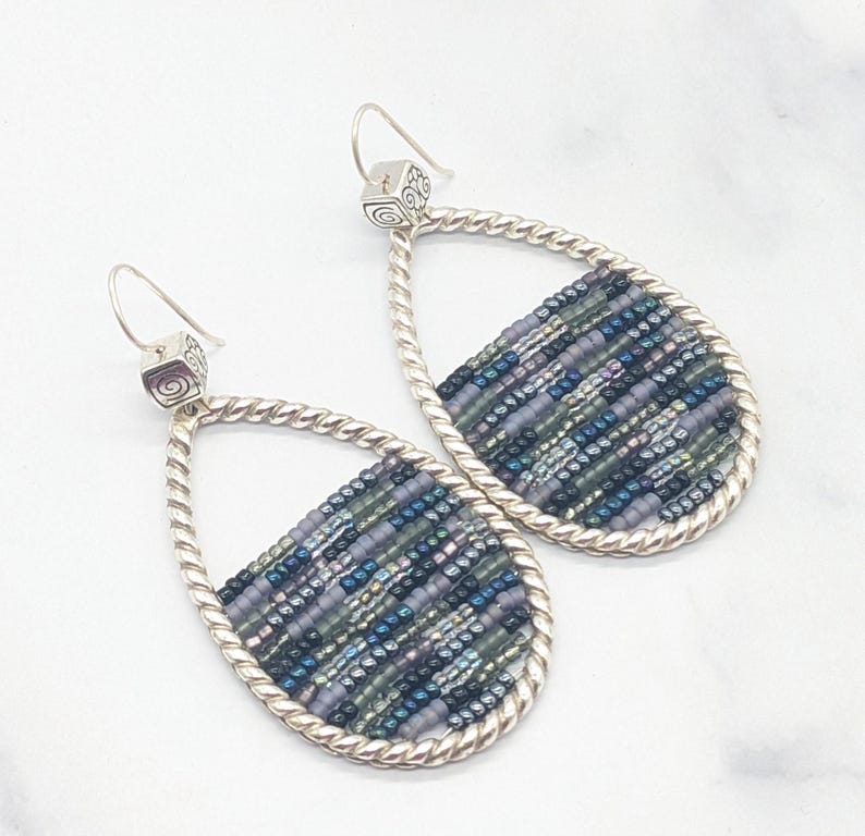May include: Teardrop-shaped earrings with a silver twisted rope design. The earrings feature a beaded section with shades of blue, green, and purple. The ear hooks have a small square design with a spiral detail. A stylish accessory.