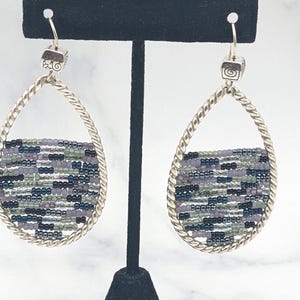 May include: Silver-toned teardrop earrings with a woven beaded design. The earrings feature a twisted metal frame and a square top with a decorative swirl. The beaded section incorporates shades of purple, green, and black.
