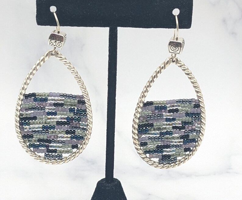 May include: Silver-toned teardrop earrings with a woven beaded design. The earrings feature a twisted metal frame and a square top with a decorative swirl. The beaded section incorporates shades of purple, green, and black.