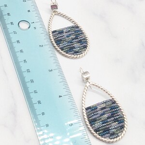 May include: A pair of teardrop earrings with a silver-tone frame and a woven bead design in shades of blue, green, and gray. The earrings are approximately 2 inches long, as measured by the ruler in the image. The earrings have a post-back closure.