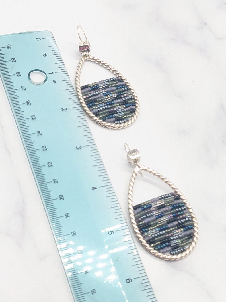 May include: A pair of teardrop earrings with a silver-tone frame and a woven bead design in shades of blue, green, and gray. The earrings are approximately 2 inches long, as measured by the ruler in the image. The earrings have a post-back closure.