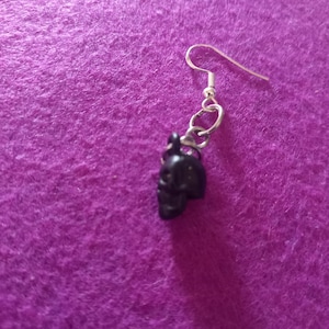 May include: A silver earring with a black skull charm. The earring is on a purple background.