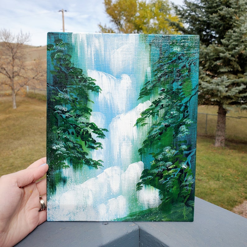 May include: A painting on canvas depicting a waterfall cascading down a cliff face, framed by lush green trees on either side. The painting is done in shades of blue, green, and white.
