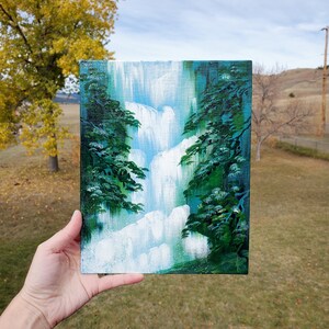 May include: A small painting on canvas depicting a waterfall cascading down a rocky cliff, surrounded by lush green trees. The painting is done in a stylized, almost cartoonish style, with bright colors and bold brushstrokes.