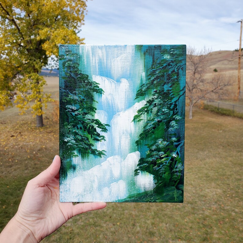 May include: A small painting on canvas depicting a waterfall cascading down a rocky cliff, surrounded by lush green trees. The painting is done in a stylized, almost cartoonish style, with bright colors and bold brushstrokes.