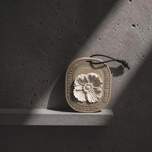 May include: An oval-shaped beige ceramic hanging ornament featuring a raised relief of a flower in the centre. The text "MAISON MANE" and "HOME DECORATION EST. 2025" is embossed along the border. A black cord is attached through a hole at the top.