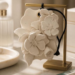 May include: A white, matte-finish ceramic plaque featuring a relief sculpture of blooming flowers and leaves. The plaque is suspended by a black cord from a gold-toned metal stand with a square base, showcasing intricate floral details.