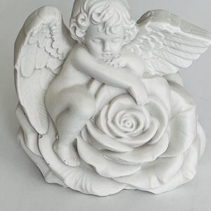 May include: A matte white figurine of a cherub with feathered wings resting its arms and head upon a large, blooming rose. The sculpture features detailed curls in the hair and layered petals on the flower, rendered in a solid, monochromatic finish.