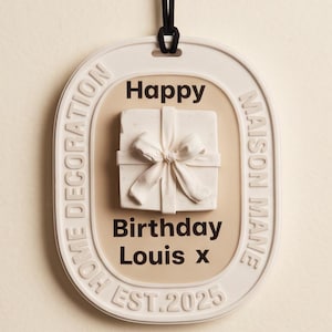 Puede incluir: Una placa colgante ovalada de color blanco roto con un cordón negro. El centro presenta una caja de regalo blanca en relieve con un lazo. El texto dice: Happy Birthday Louis x, HOME DECORATION, MAISON MANE y EST. 2025. El fondo es de color beige claro.