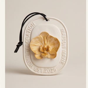 May include: An oval-shaped white ceramic hanging plaque featuring a raised, textured golden-yellow orchid flower in the center. The border includes the embossed text MAISON MANE, HOME DEFORATION, and EST. 2025, with a black cord attached at the top.