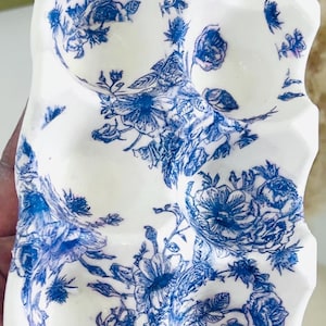 May include: A white, ceramic decorative piece with a blue floral pattern. The design features detailed blue flowers and foliage against a white background. The edges of the piece are uneven, giving it a unique shape.