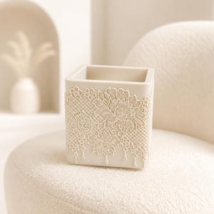 May include: A square, off-white container featuring a raised, intricate lace-like floral pattern on its exterior. The design includes detailed mesh textures and delicate hanging elements, creating a textured surface on the solid, light-coloured vessel.